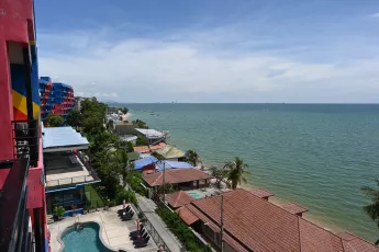 DZ6_1318: Seaside view from a colorful beachfront balcony overlooking rooftops, palm trees, a swimming pool, and the calm blue ocean stretching to the horizon.