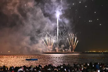 DFC_3850: A crowd gathers along the shore as fireworks burst and reflect across the water, lighting up the night sky and sending plumes of smoke into the air.