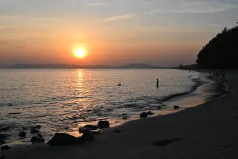 DFC_3873: A tranquil sunset over a quiet beach, the sun casting a golden path across calm waters as silhouettes of swimmers and trees line the shoreline.