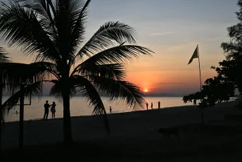 DFC_3882: Silhouetted palm trees and beachgoers frame a fiery sun sinking into the calm ocean at sunset.