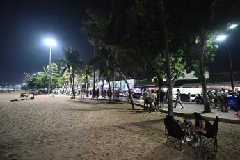 DZ6_1724: Nighttime beach promenade lined with palm trees and streetlights, people strolling and gathering along the sand.