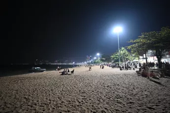 DZ6_1721: Nighttime beach scene, soft sand, glowing streetlights, and people strolling and relaxing along the shoreline under a starless sky.