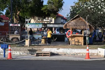 DZ6_1833: A small waterfront stall with a thatched-roof hut and colorful life ring, a person in a yellow shirt standing nearby, and trees and houses in the background on a sunny day.
