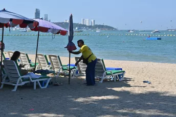 DFC_4033: A beach attendant adjusts a sun umbrella beside empty lounge chairs as calm blue water and distant city buildings stretch along the shoreline.