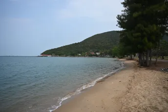 DFC_4446: Quiet shoreline with gentle waves lapping the sand and a tree-lined coast stretching toward a green hillside.