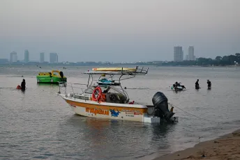 DFC_5305: Evening calm at Klet Kaeo, a small motorboat rests near the shore as swimmers and distant city towers fade into the twilight.